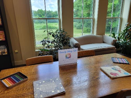 A wooden table with coloring books and colored pencils sits by large windows overlooking a green lawn. A white couch and potted plants are nearby, creating a cozy, inviting space for creative activities.